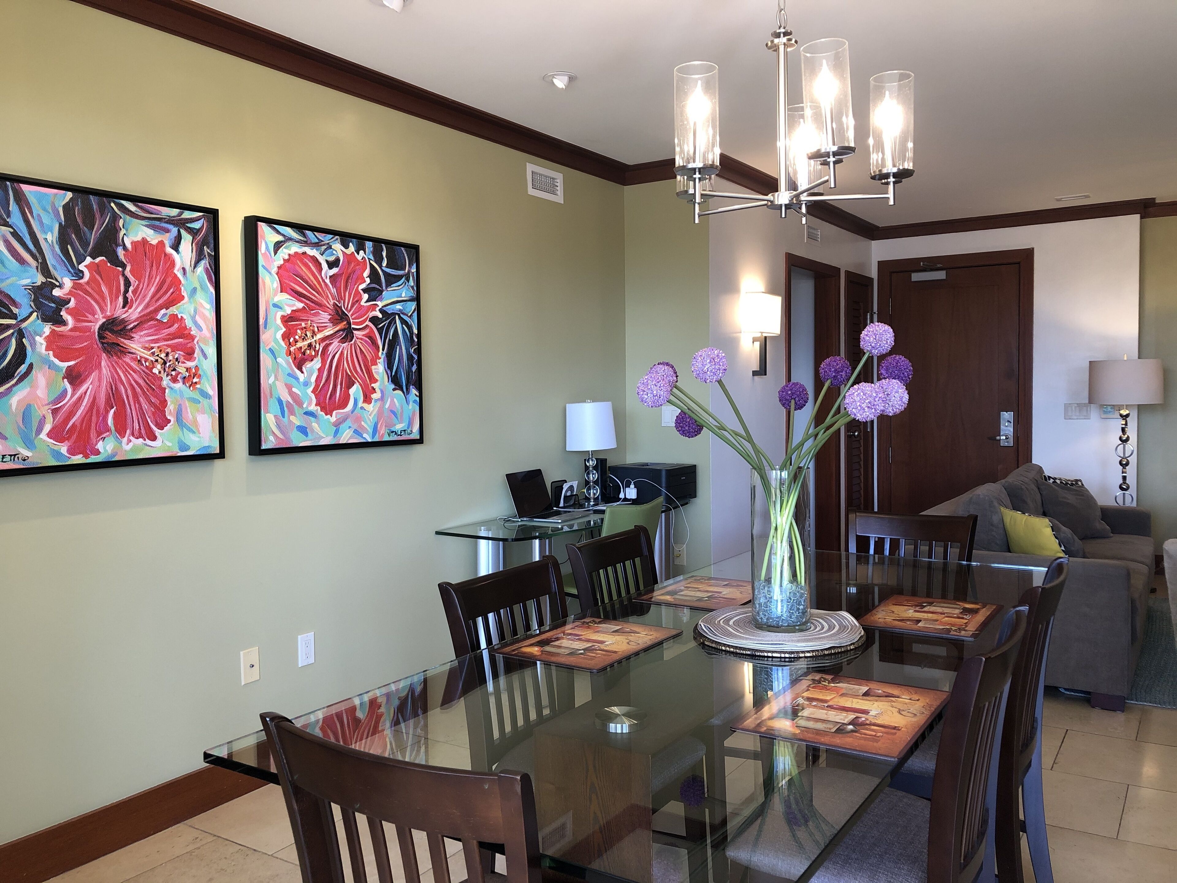 Un coin repas avec une table en verre, des chaises et un vase rempli de fleurs violettes. Deux grandes peintures représentant des motifs floraux ornent le mur.