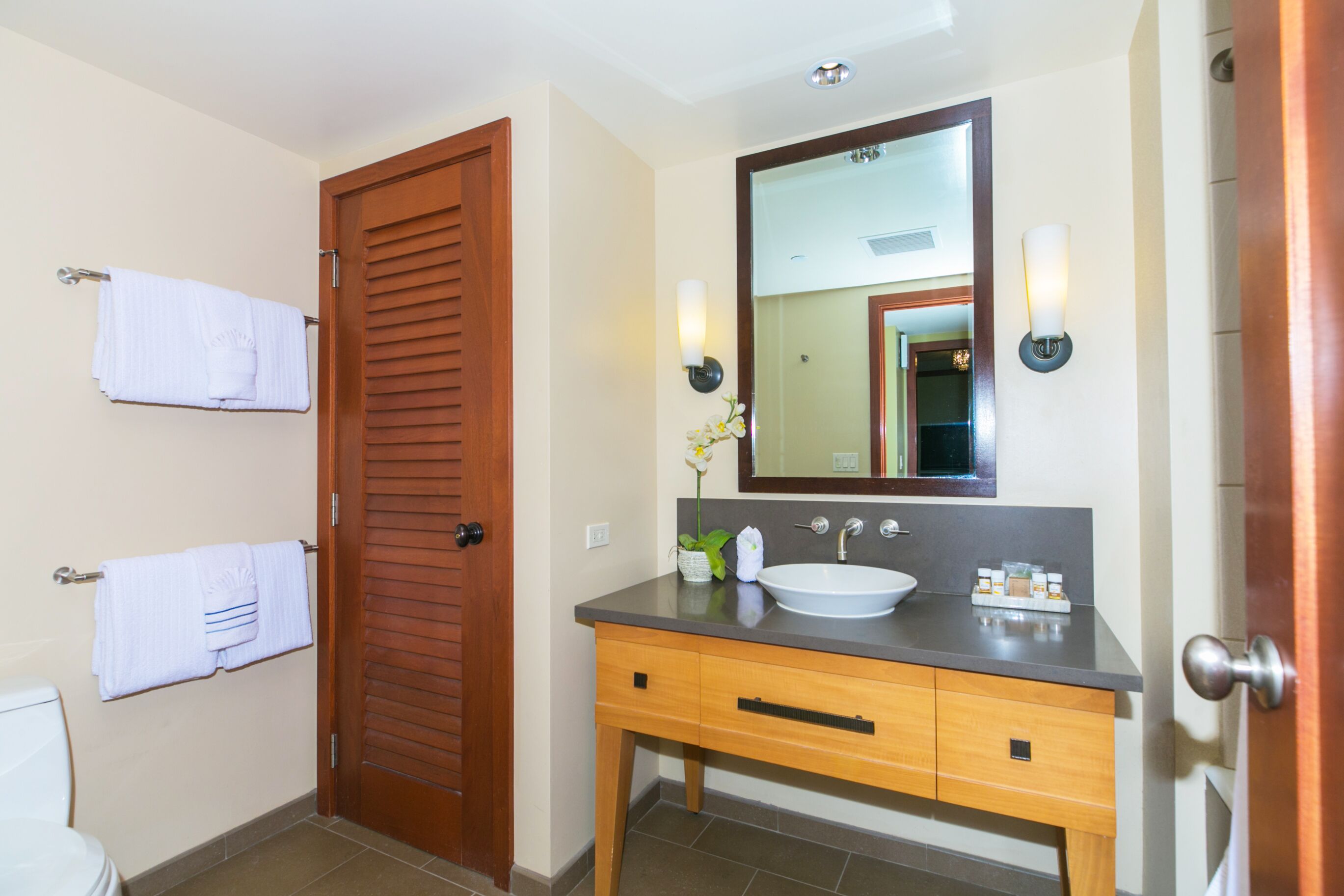 Un baño con puerta de madera, un lavabo blanco, un espejo y dos luces empotradas.