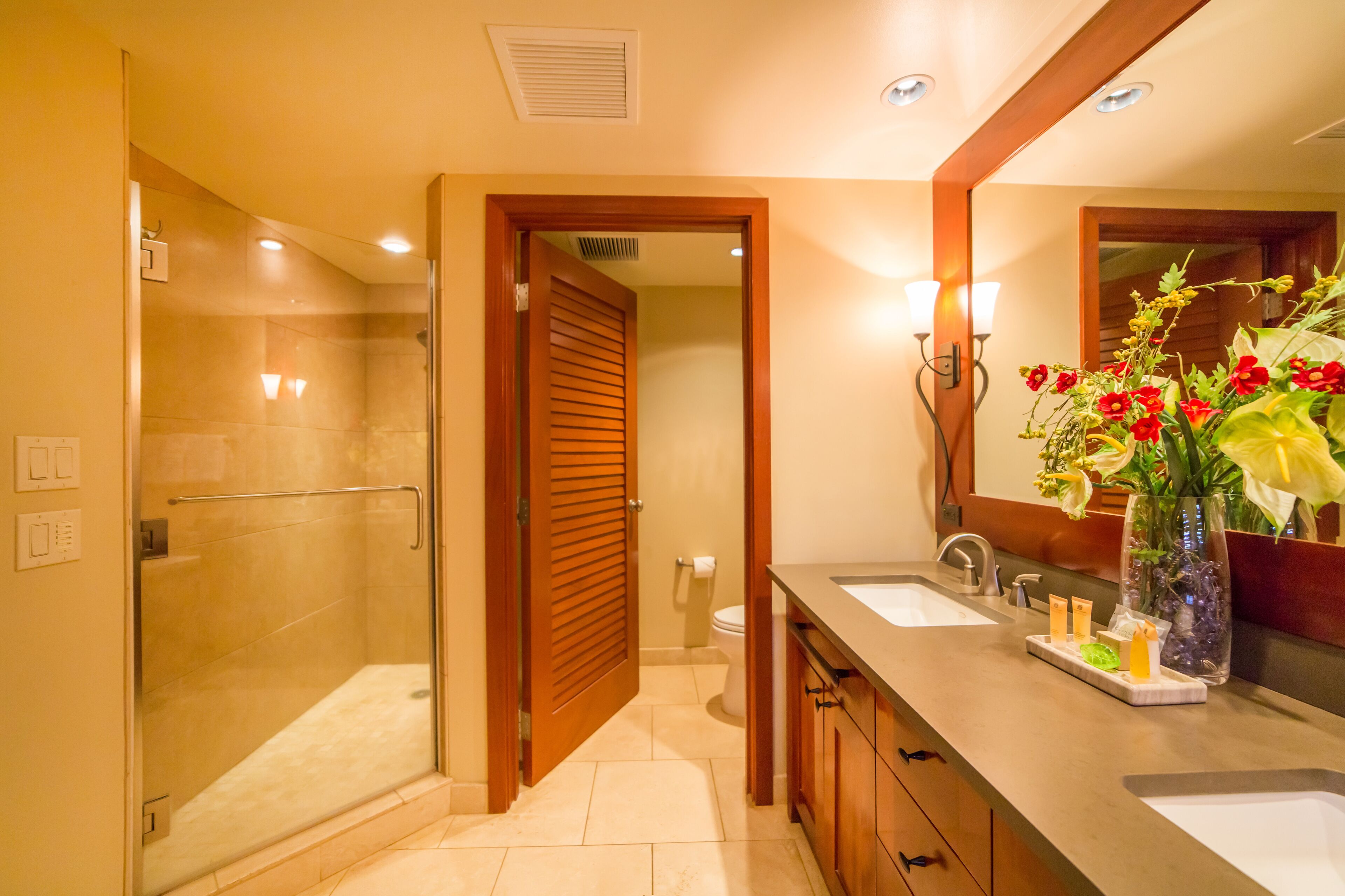 Un baño moderno con ducha envuelta en vidrio, puerta de madera, tocador con espejo y un jarrón de flores.