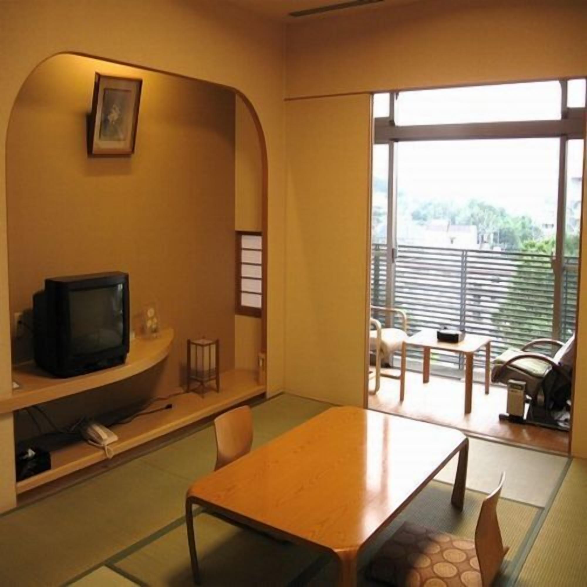 A room with a television on a shelf, a table, chairs, and a view through a large window.