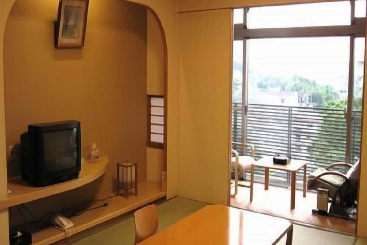 A room with a television on a shelf, a table, chairs, and a view through a large window.