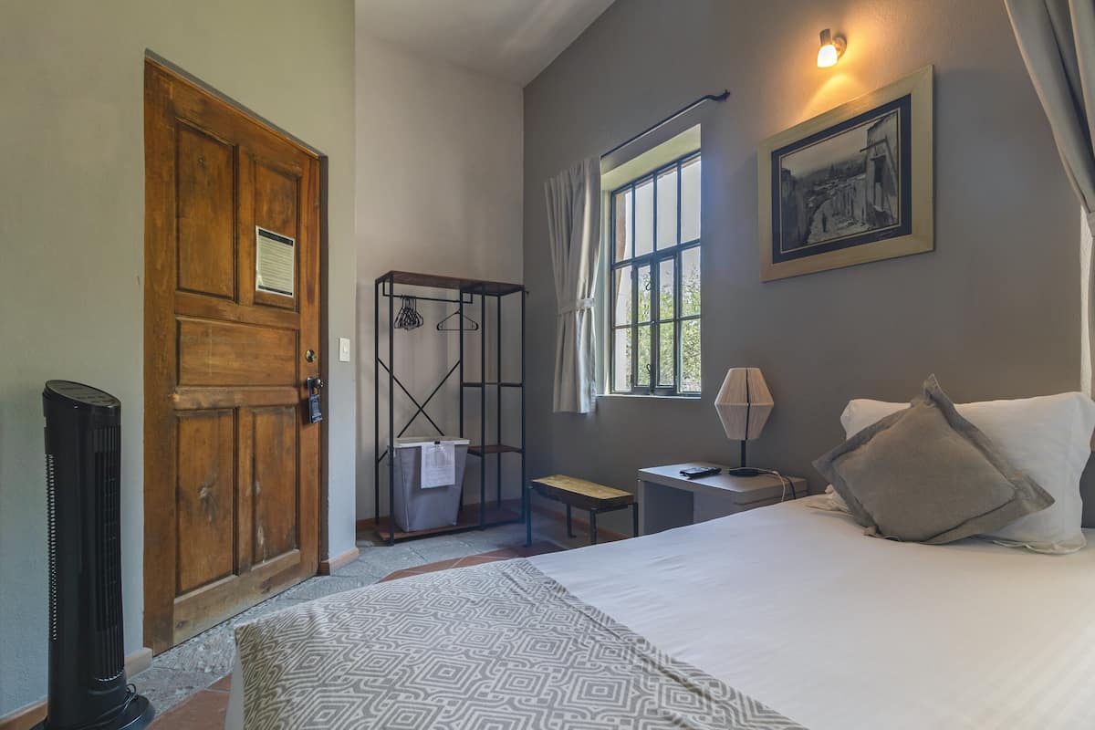 A bedroom with a large bed, a wooden door, a window with curtains, a bedside table, and a wall-mounted picture.