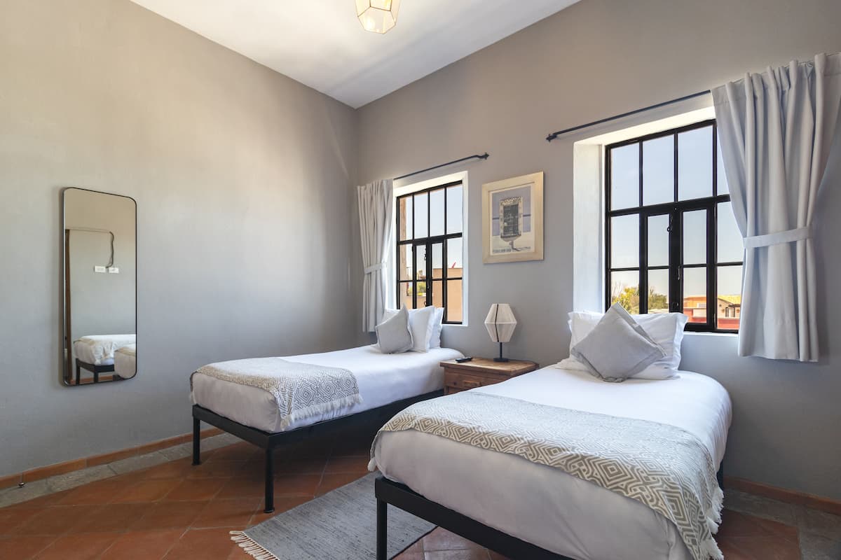 A bedroom with two beds, a large window with curtains, a mirror, and a framed picture on the wall.