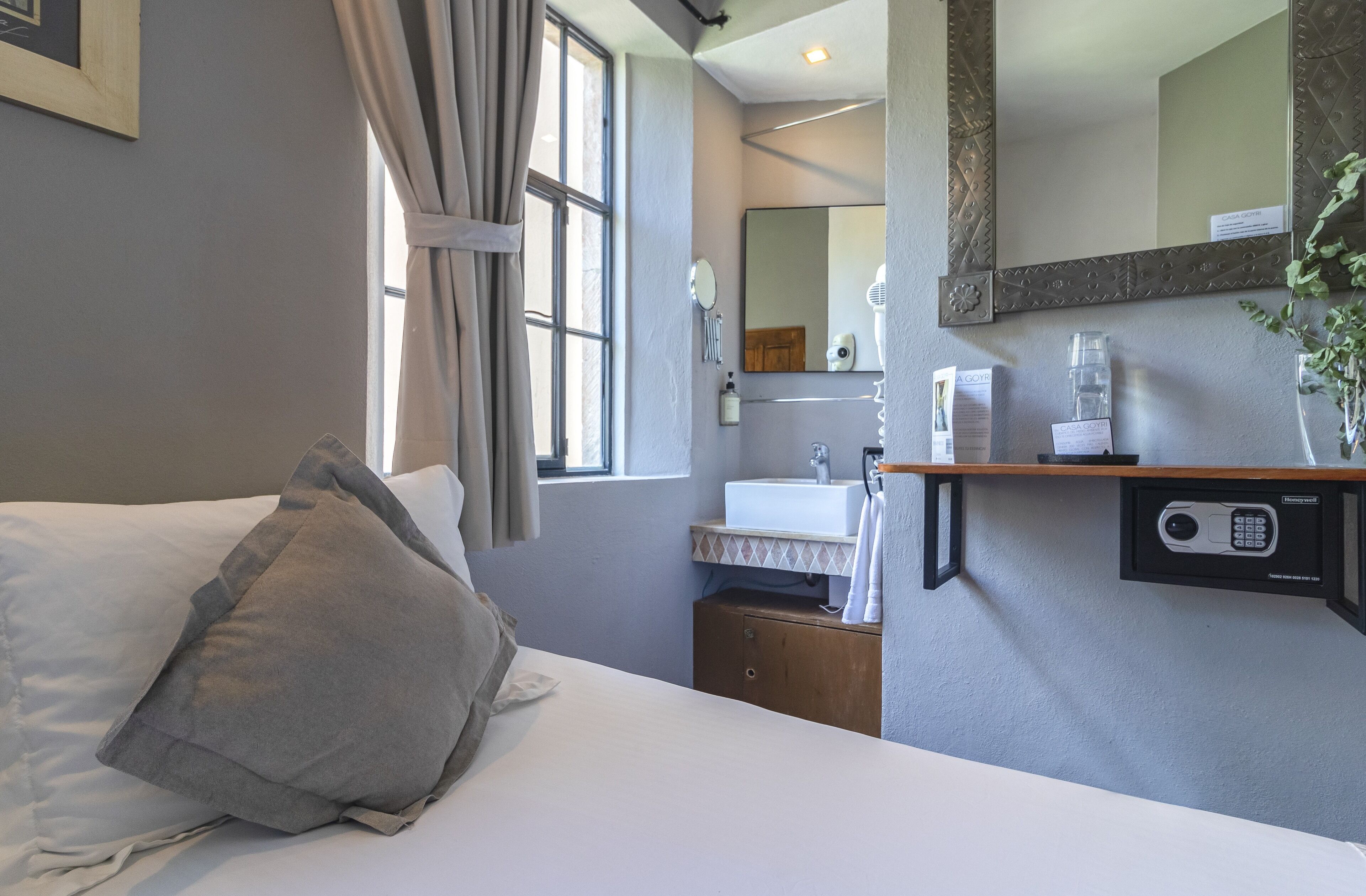 Ein Hotelzimmer mit Bett, Fenster mit Vorhängen, Bad mit Waschbecken und Spiegel sowie einem Sicherheitstastenfeld an der Wand.