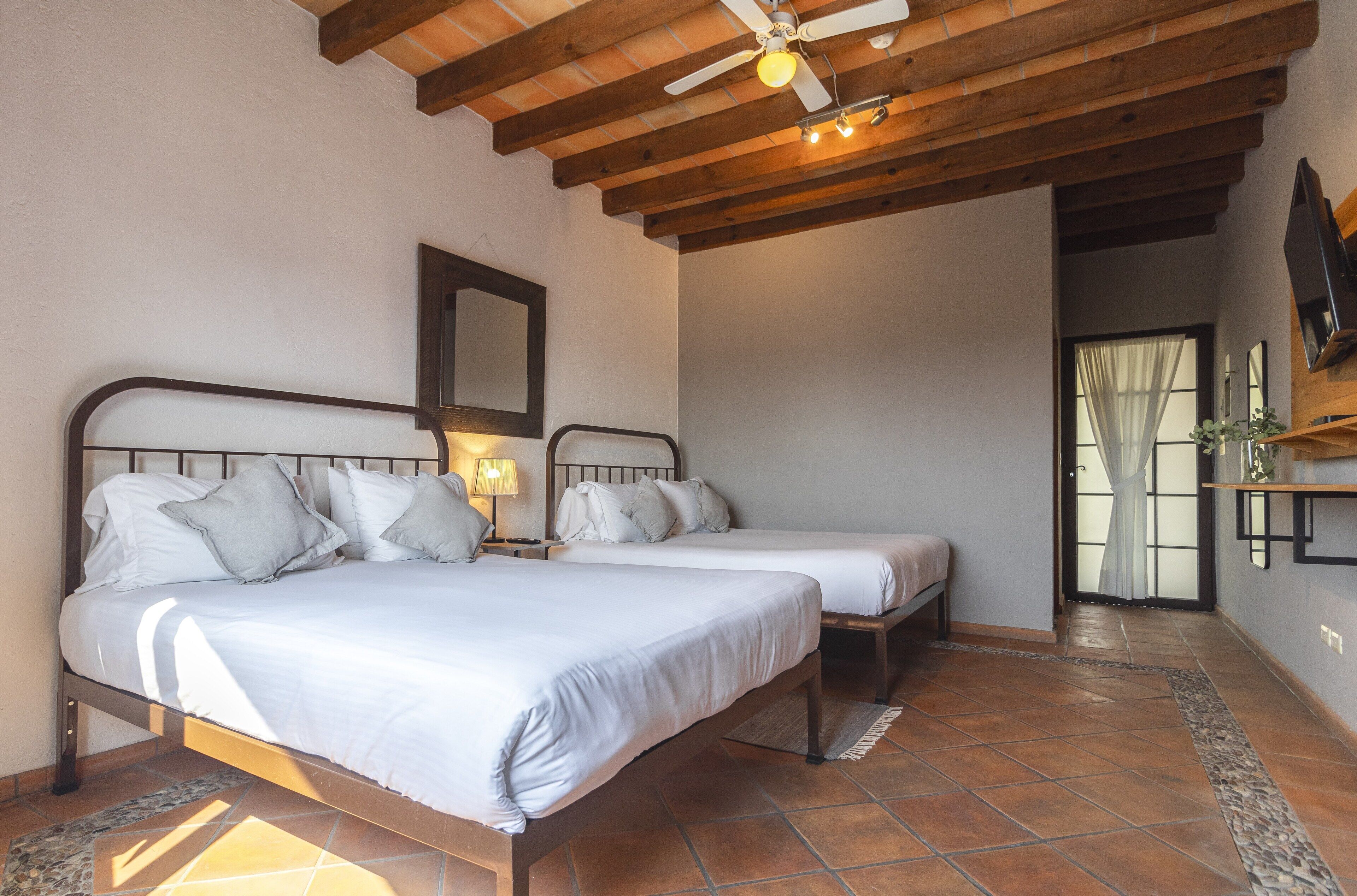 A bedroom with two beds, wooden beams, a ceiling fan, a nightstand with a lamp, and a TV on a shelf.