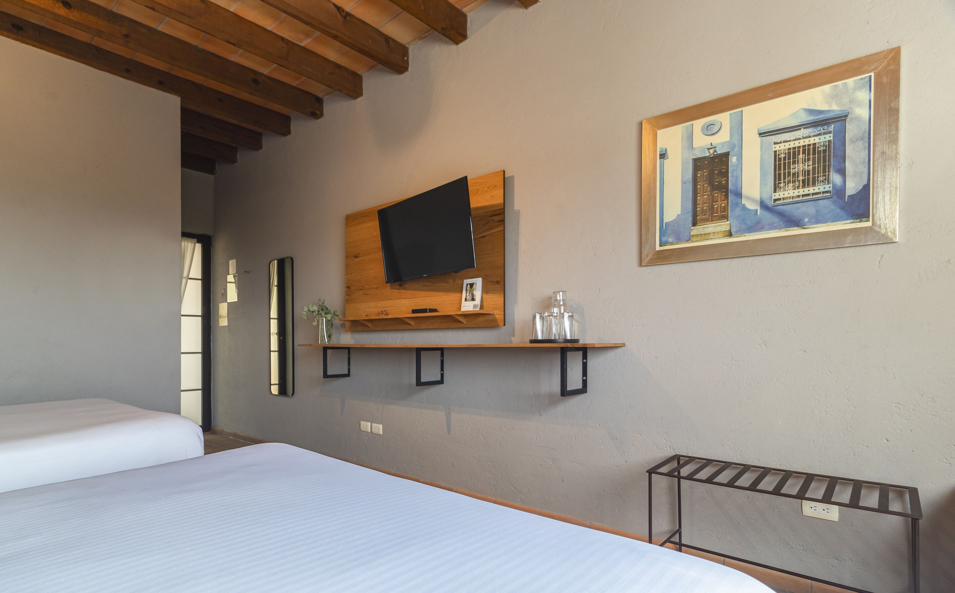 A hotel room with a bed, a mounted TV, a painting, and a small shelf with items.