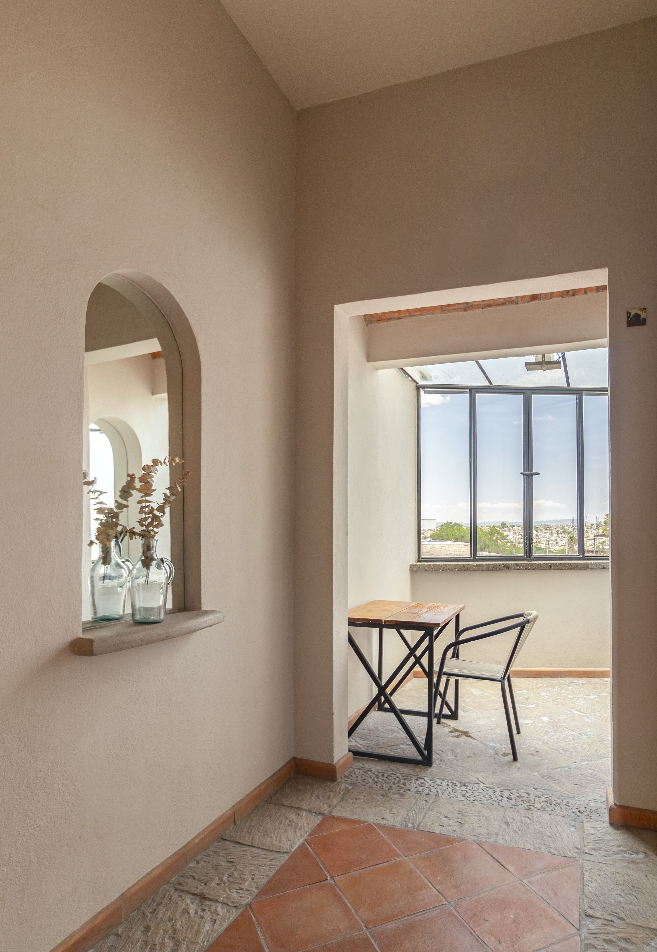 A corridor with a tiled floor, a small table with chairs, a window with a view, and a mirror reflecting a vase with flowers.