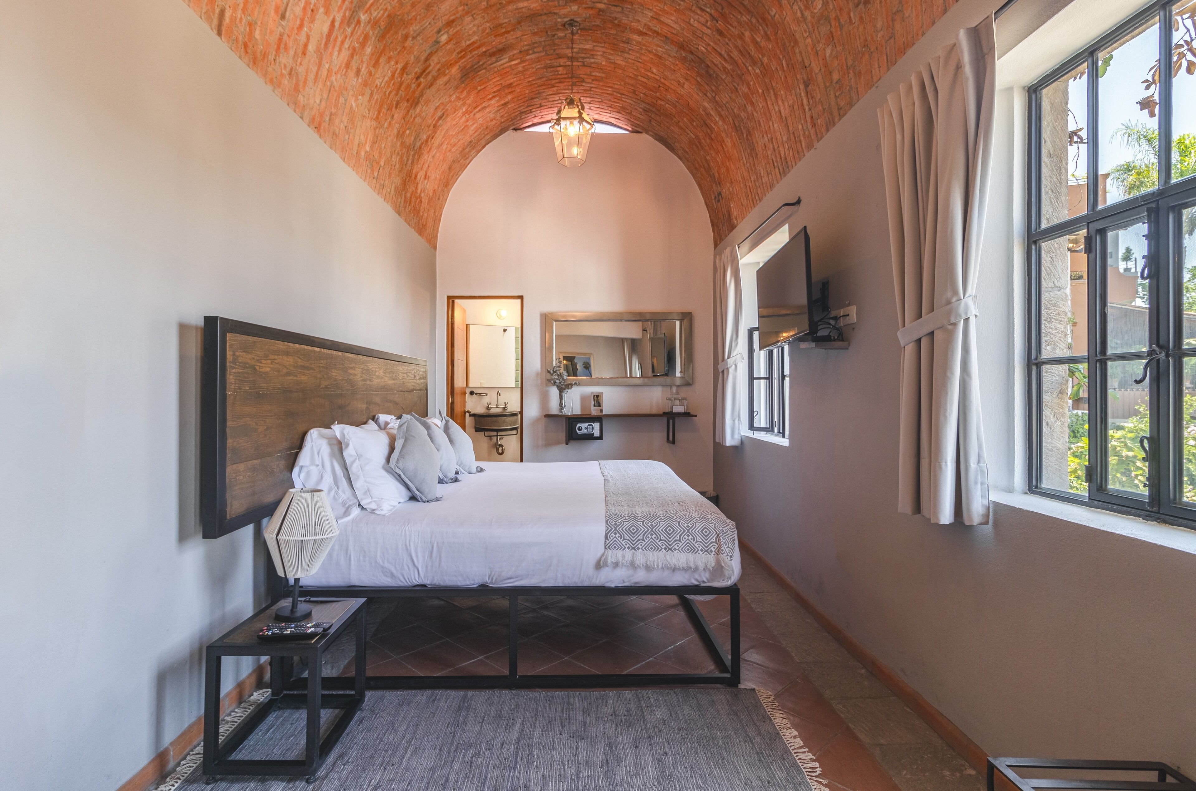 Un dormitorio con techo de ladrillo, una cama grande con ropa blanca, una mesita de noche, un tocador, un espejo y una ventana con cortinas.