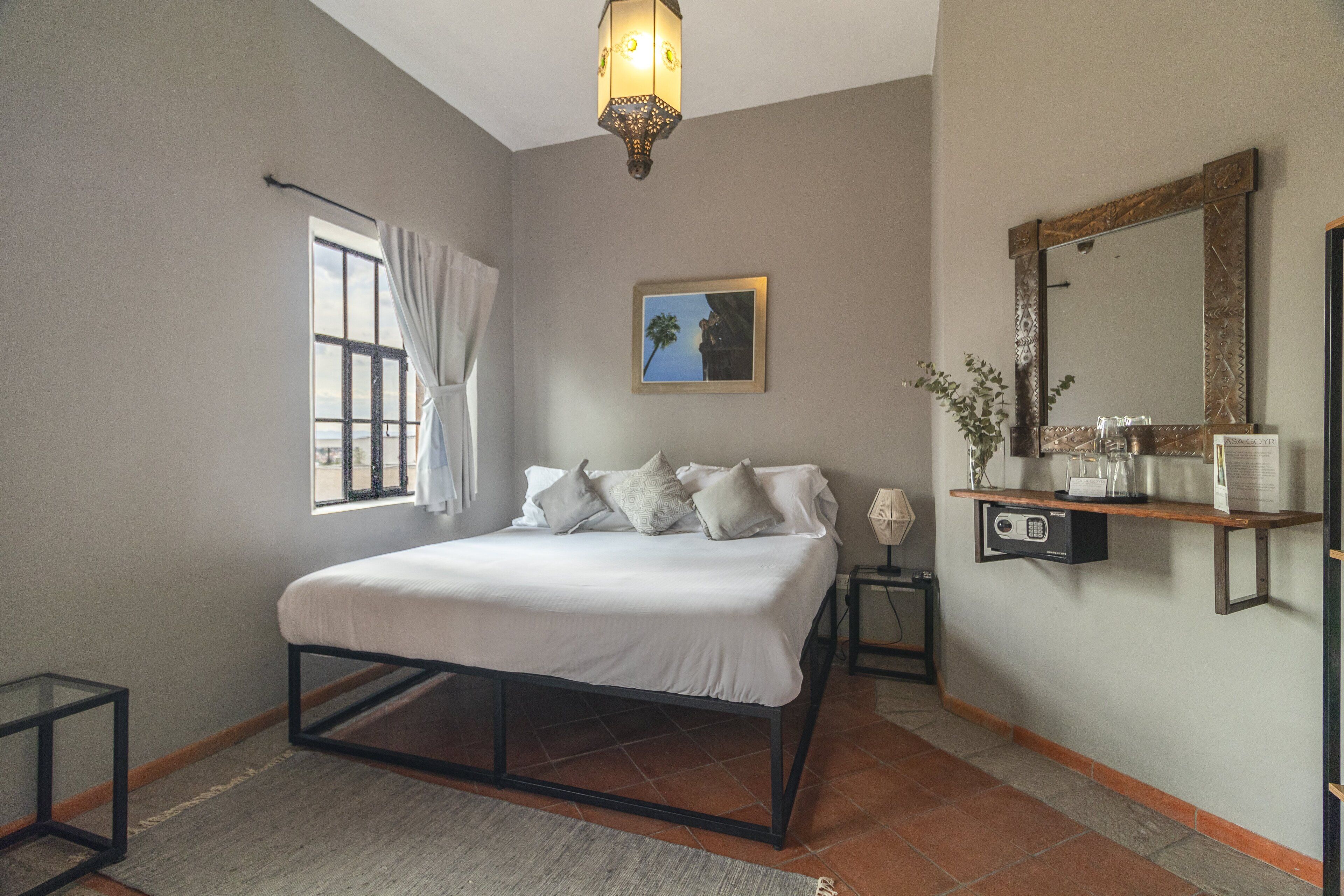 Une chambre à coucher avec un lit, une fenêtre avec des rideaux, une étagère fixée au mur avec des articles de toilette et un luminaire au plafond.