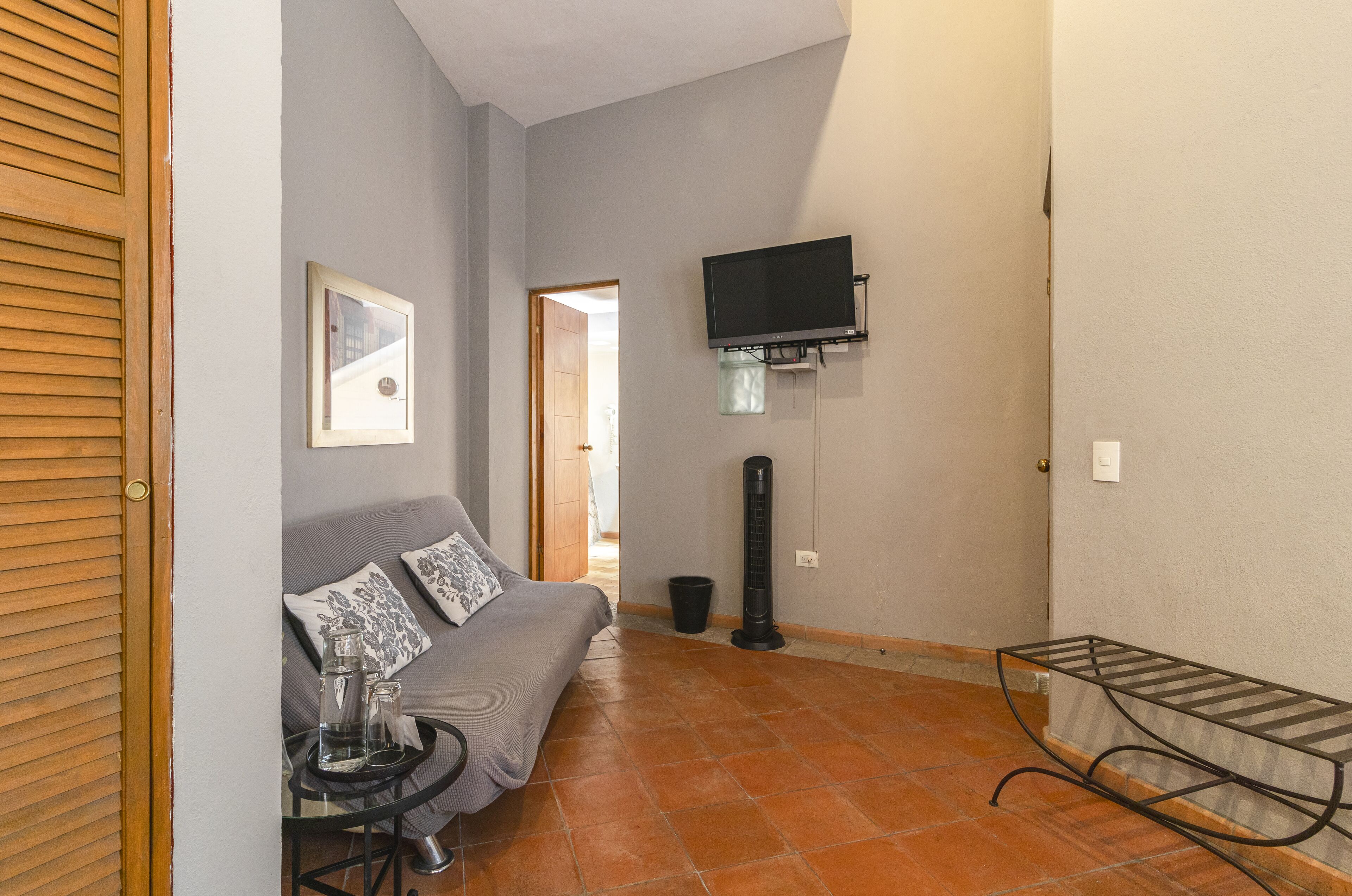 A small living room with a grey sofa, a coffee table, a TV mounted on the wall, and a tiled floor.