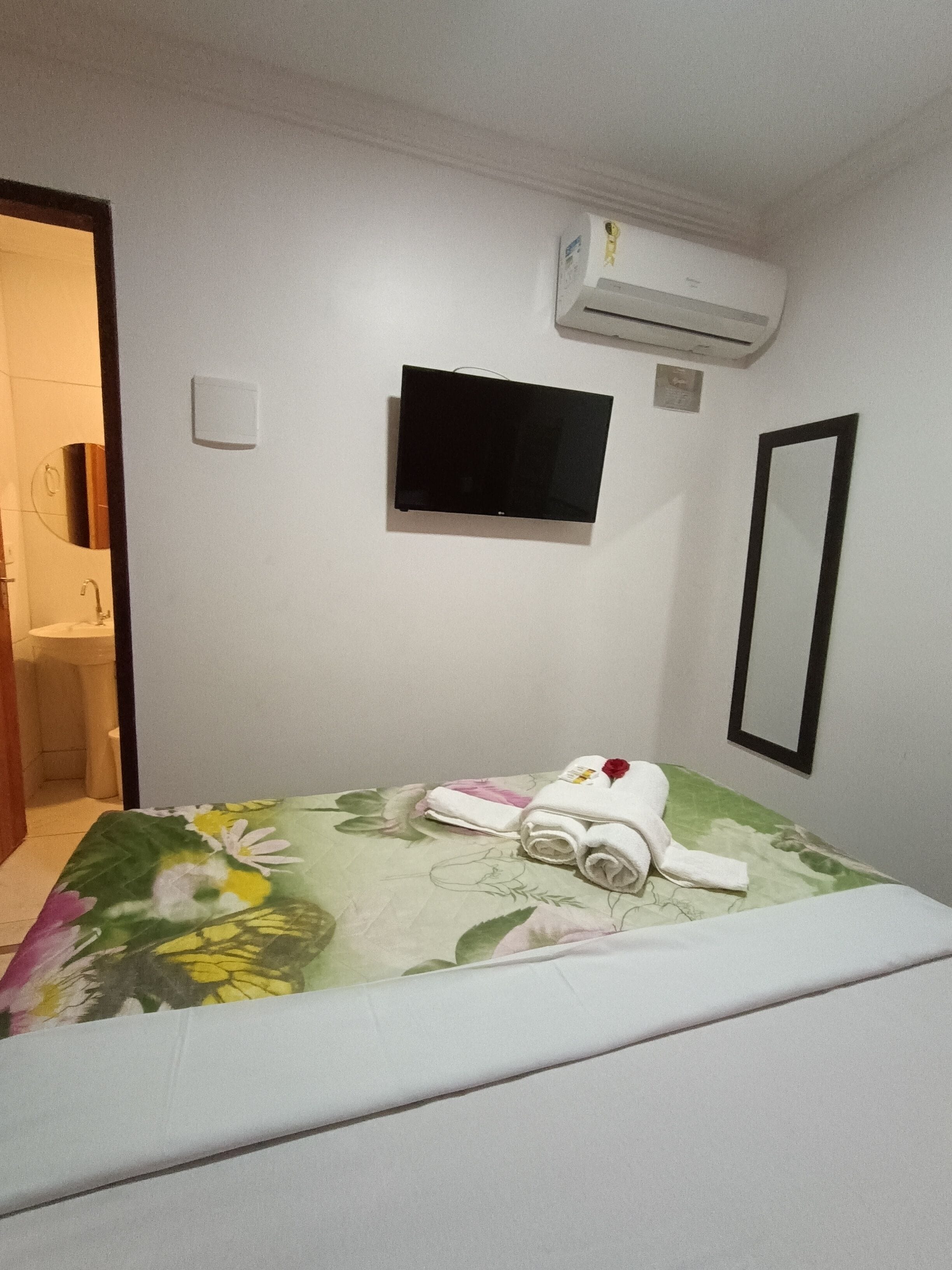Habitación de hotel con cama, televisión, aire acondicionado, espejo y baño visible a través de una puerta abierta.