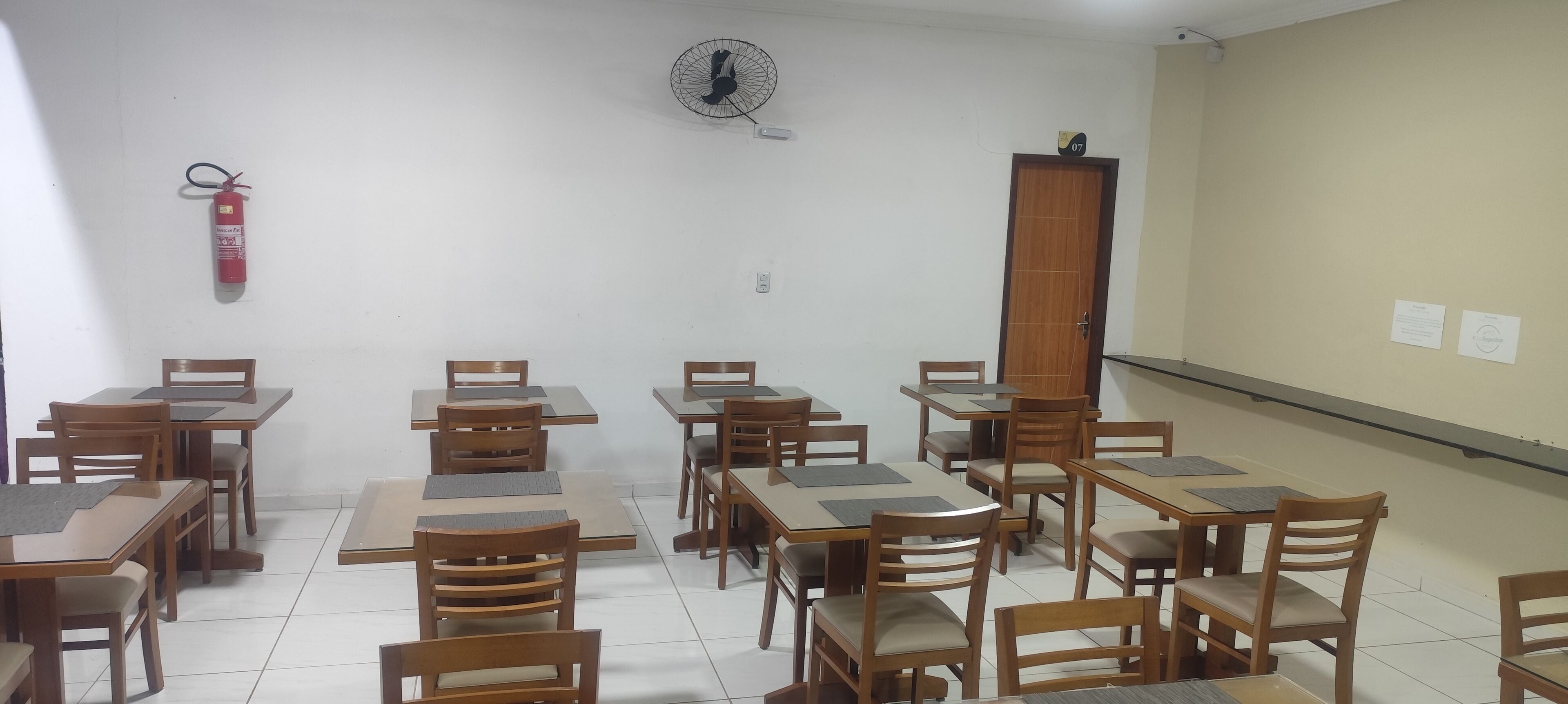 Un aula con mesas y sillas de madera, un extintor, un ventilador y una puerta con el número 07.