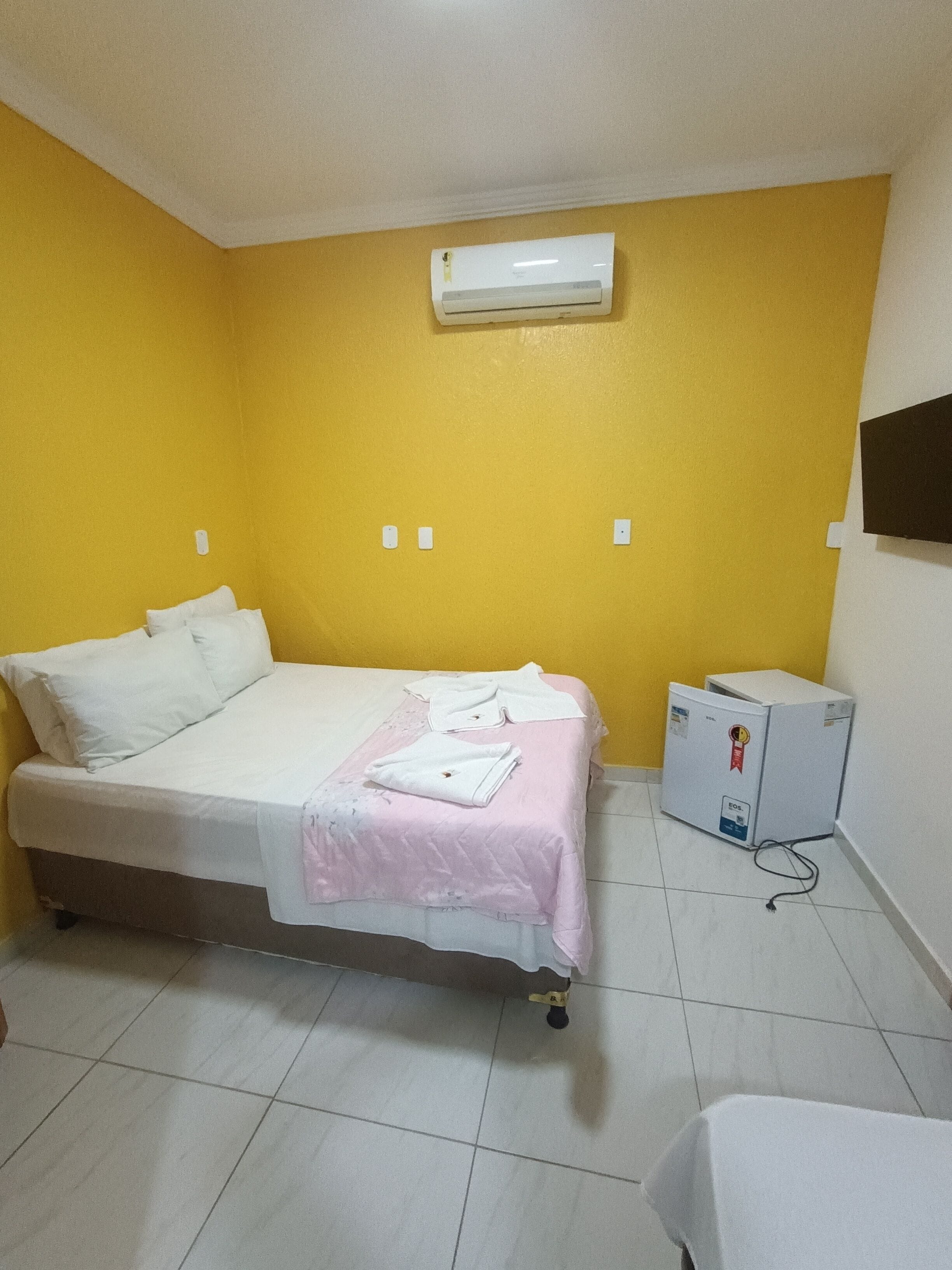 A room with a bed, a small refrigerator, and an air conditioning unit on the wall.