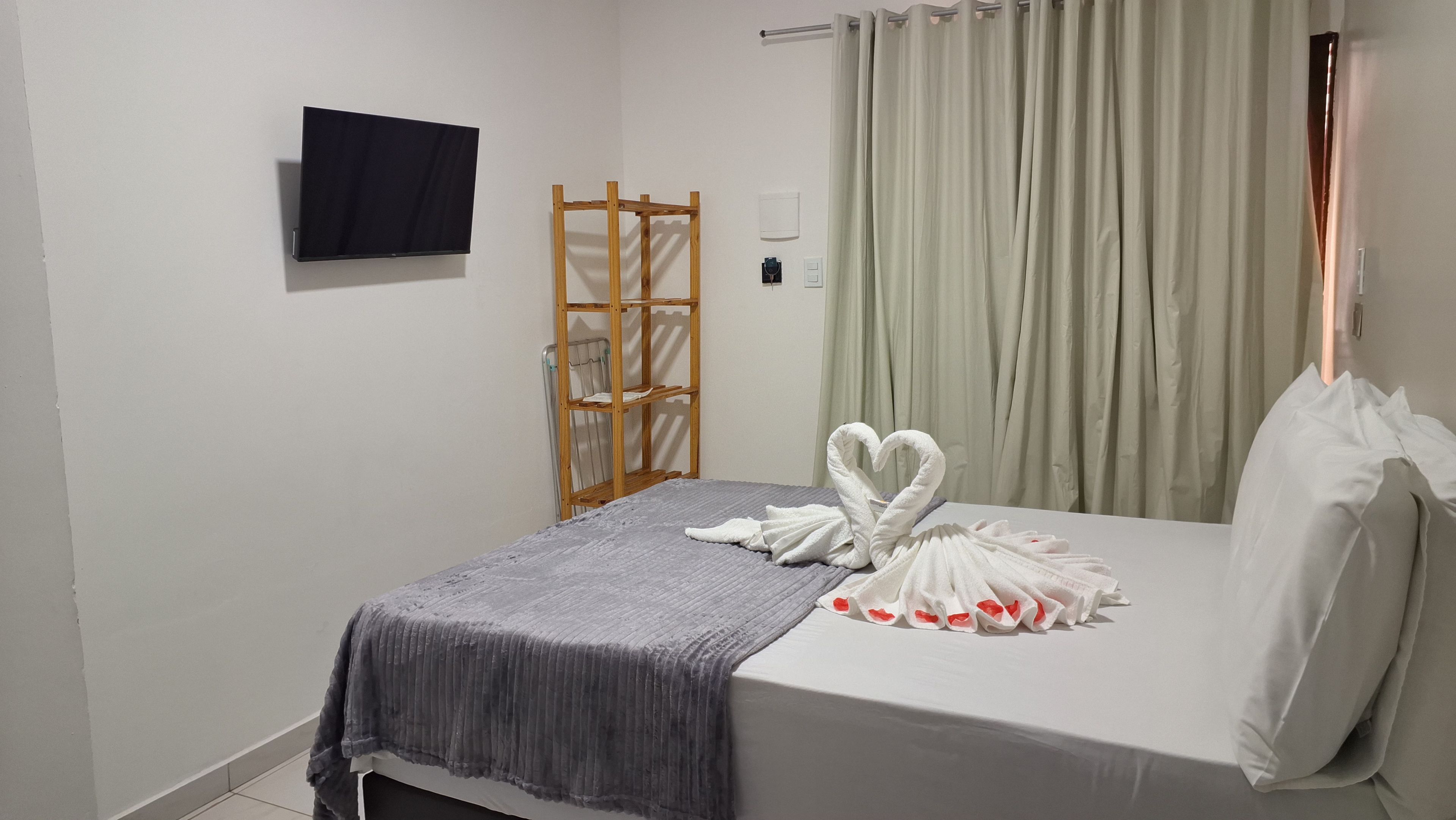Un lit simple avec une couverture grise, une étagère en bois, un téléviseur à écran plat fixé au mur et un rideau.