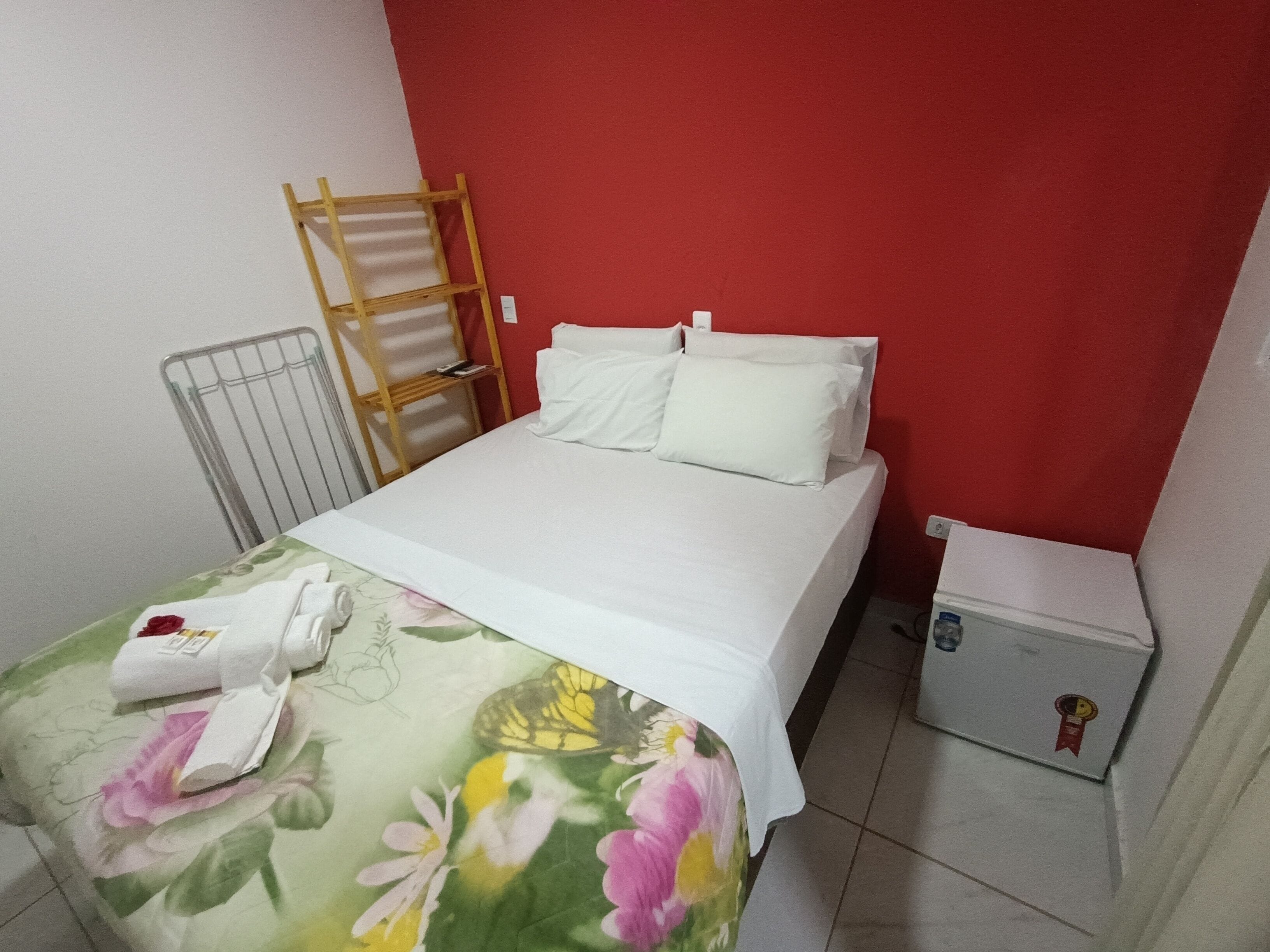 A neatly made bed with white linens, a floral bedspread, a wooden ladder against the wall, a metal radiator, and a small bedside table with a red plaque.