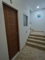 Una escalera con barandilla, una puerta con un cartel de número y una ventana pequeña con cortina.