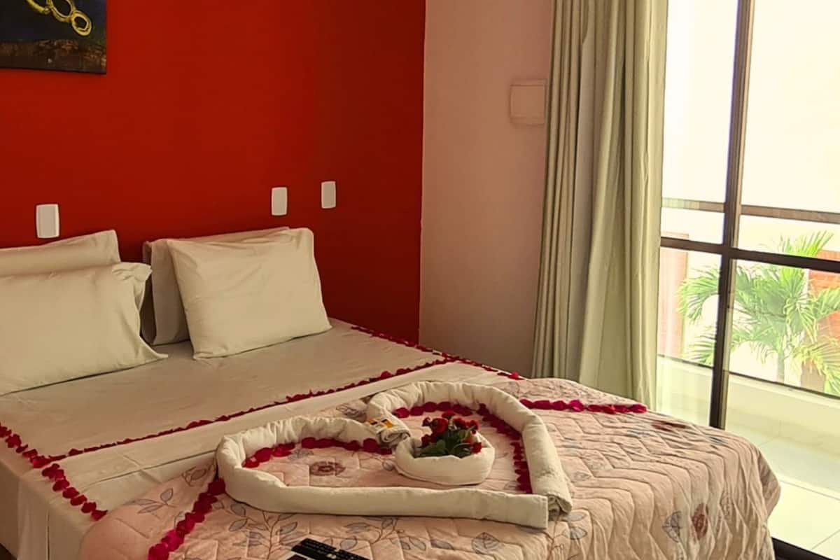 Ein Hotelzimmer mit einem großen Bett, einer roten Akzentwand, einem Fenster mit Vorhängen und Blick auf Palmen draußen.