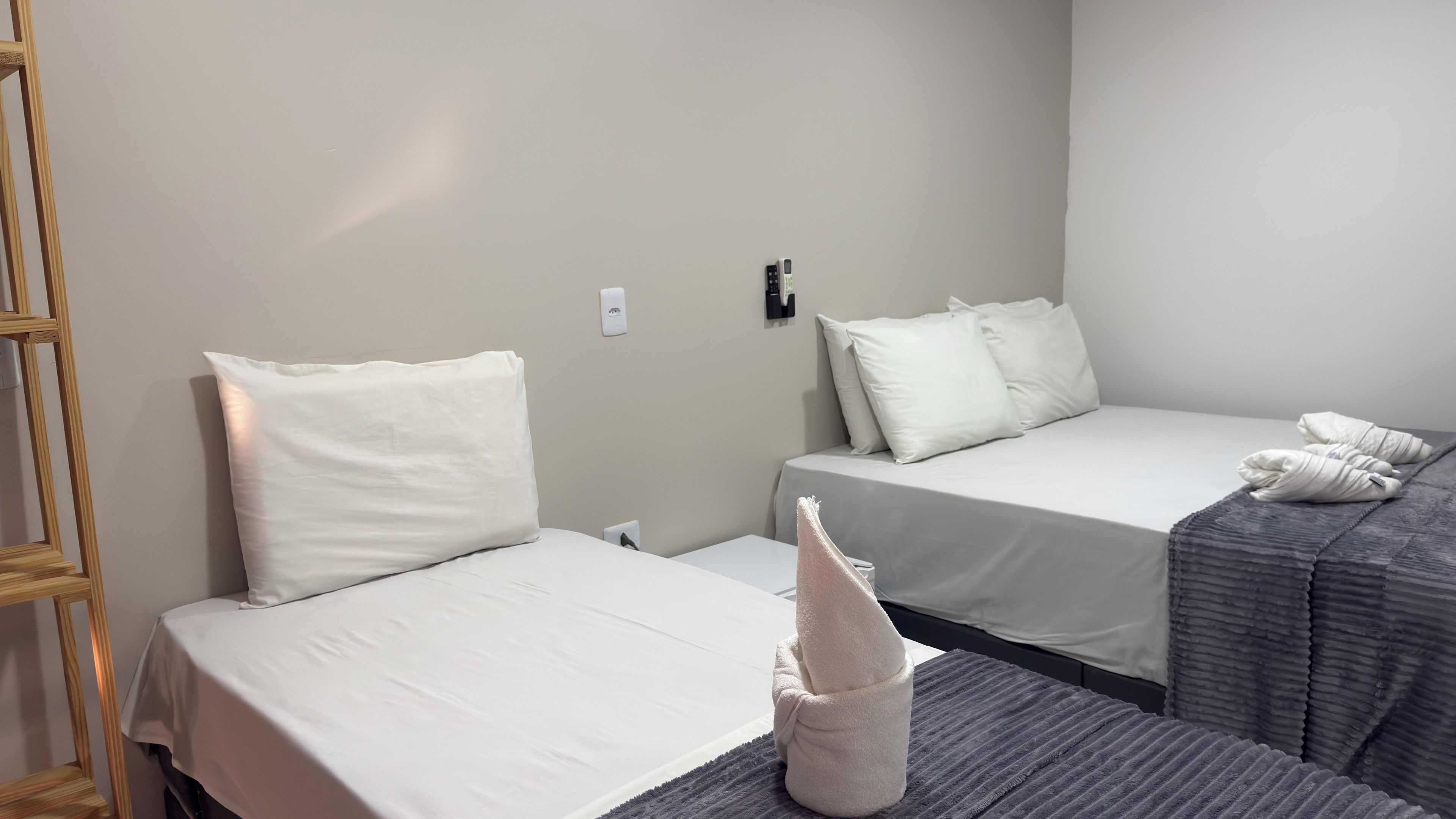 A hotel room with two single beds, a wooden ladder, a towel, and a wall-mounted light switch.