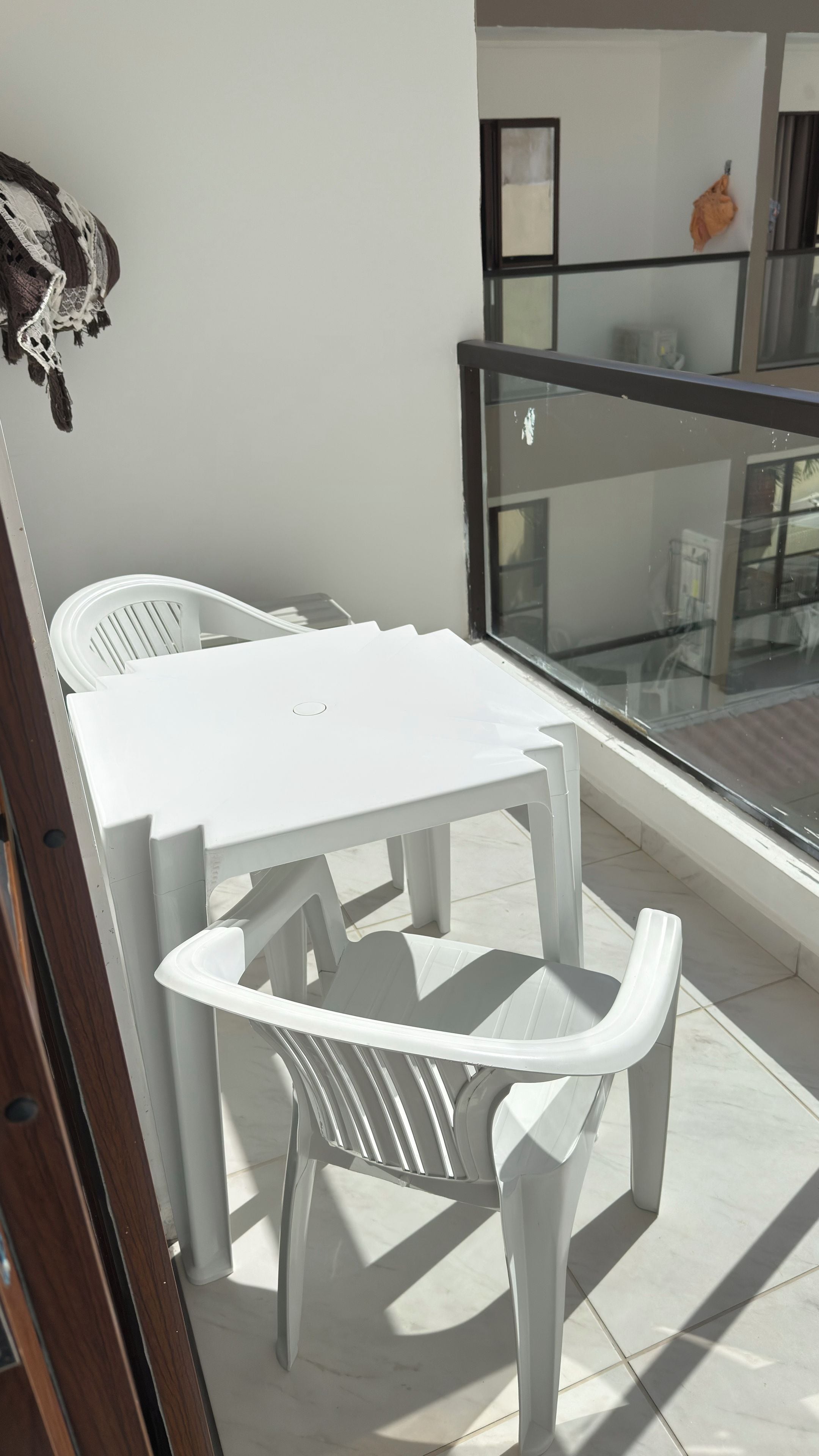 Une table et des chaises en plastique blanc sur un balcon doté d’une balustrade en verre, avec des murs blancs et une fenêtre.