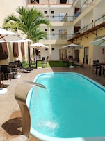 Una piscina con sillones de descanso y sombrillas, rodeada por un edificio de varios pisos con balcones.