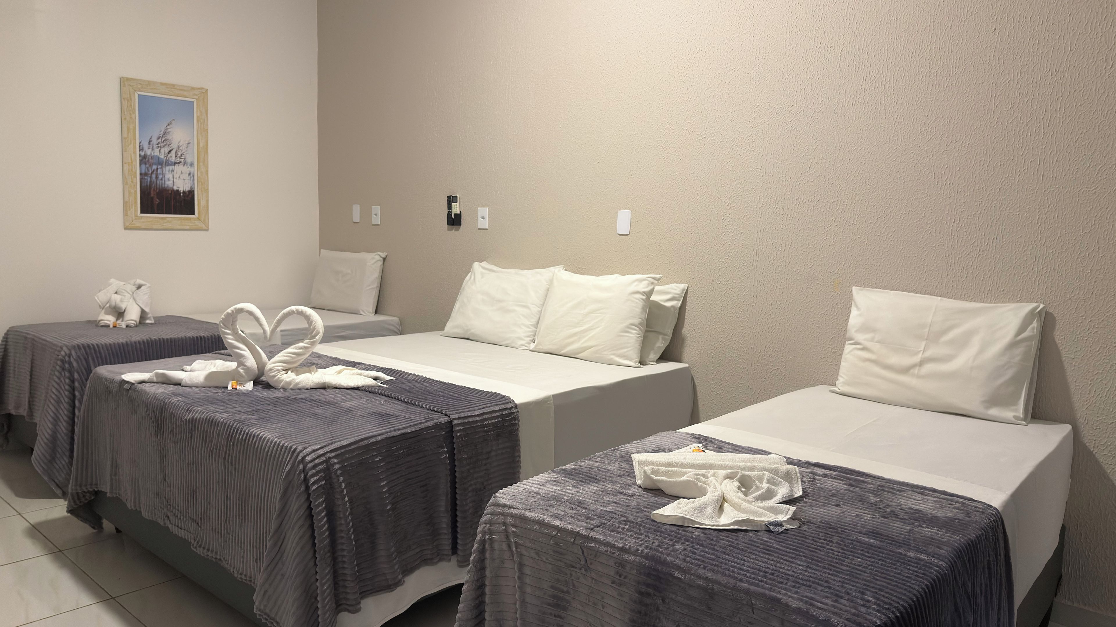 Ein Hotelzimmer mit zwei Einzelbetten, jeweils mit grauer Bettdecke und weißen Kissen. An der Wand hängt ein gerahmtes Bild und ein kleiner Tisch mit Handtüchern.