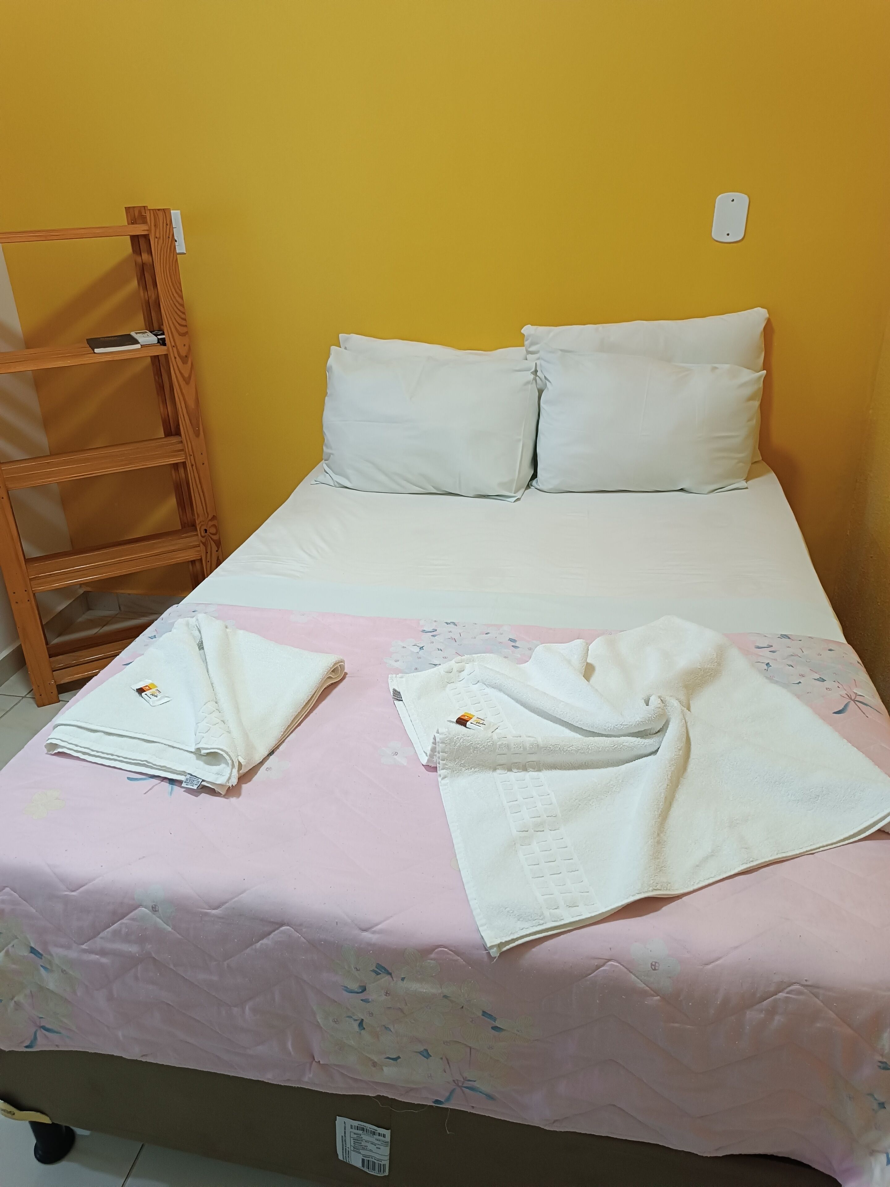 Uma cama de solteiro com roupa de cama e almofadas brancas, uma escada de madeira e um edredom rosa com estampas florais.