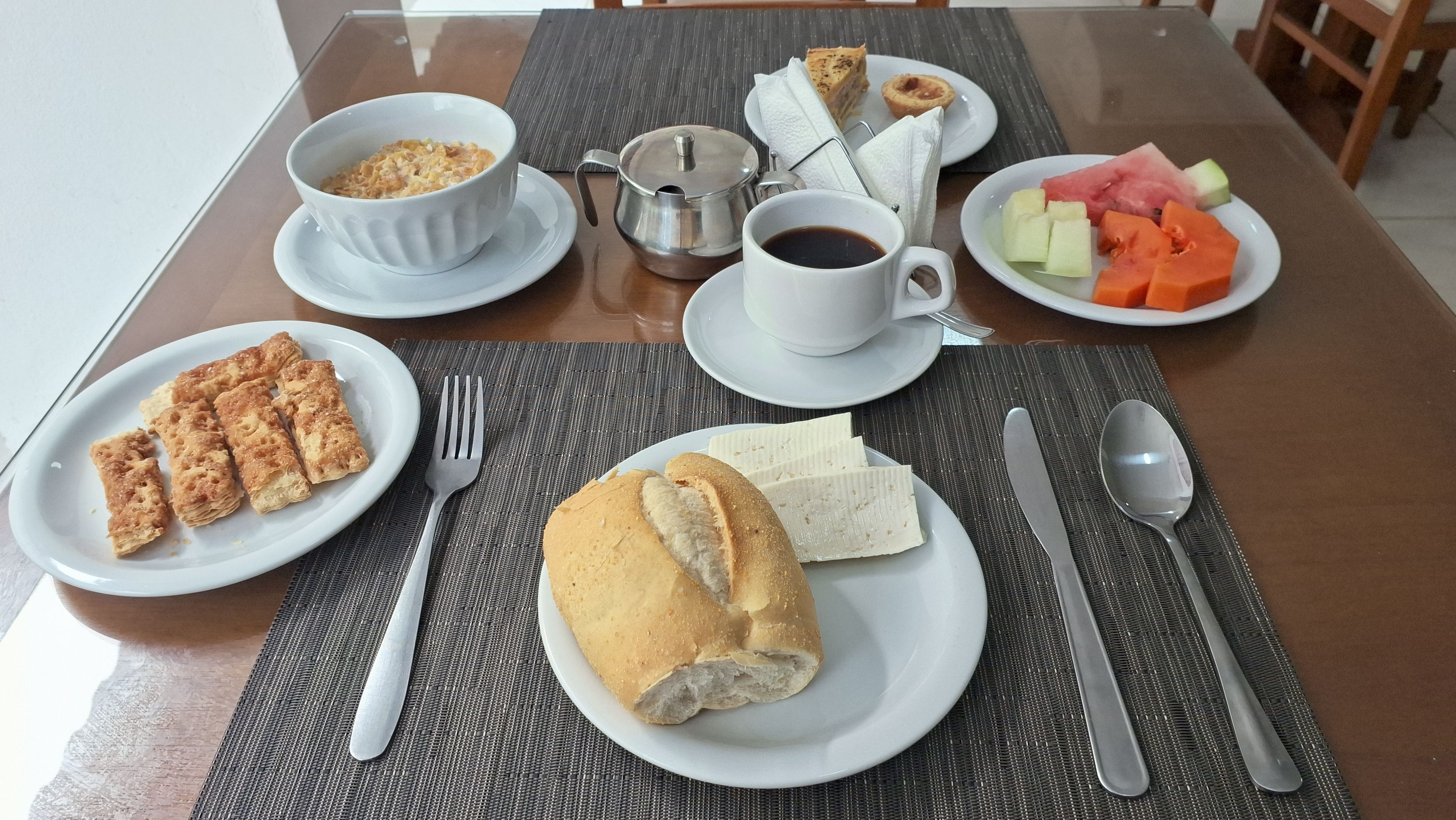 A breakfast setting with a cup of coffee, a bowl of cereal, a plate of pastries, a sandwich, and a plate of fruit.