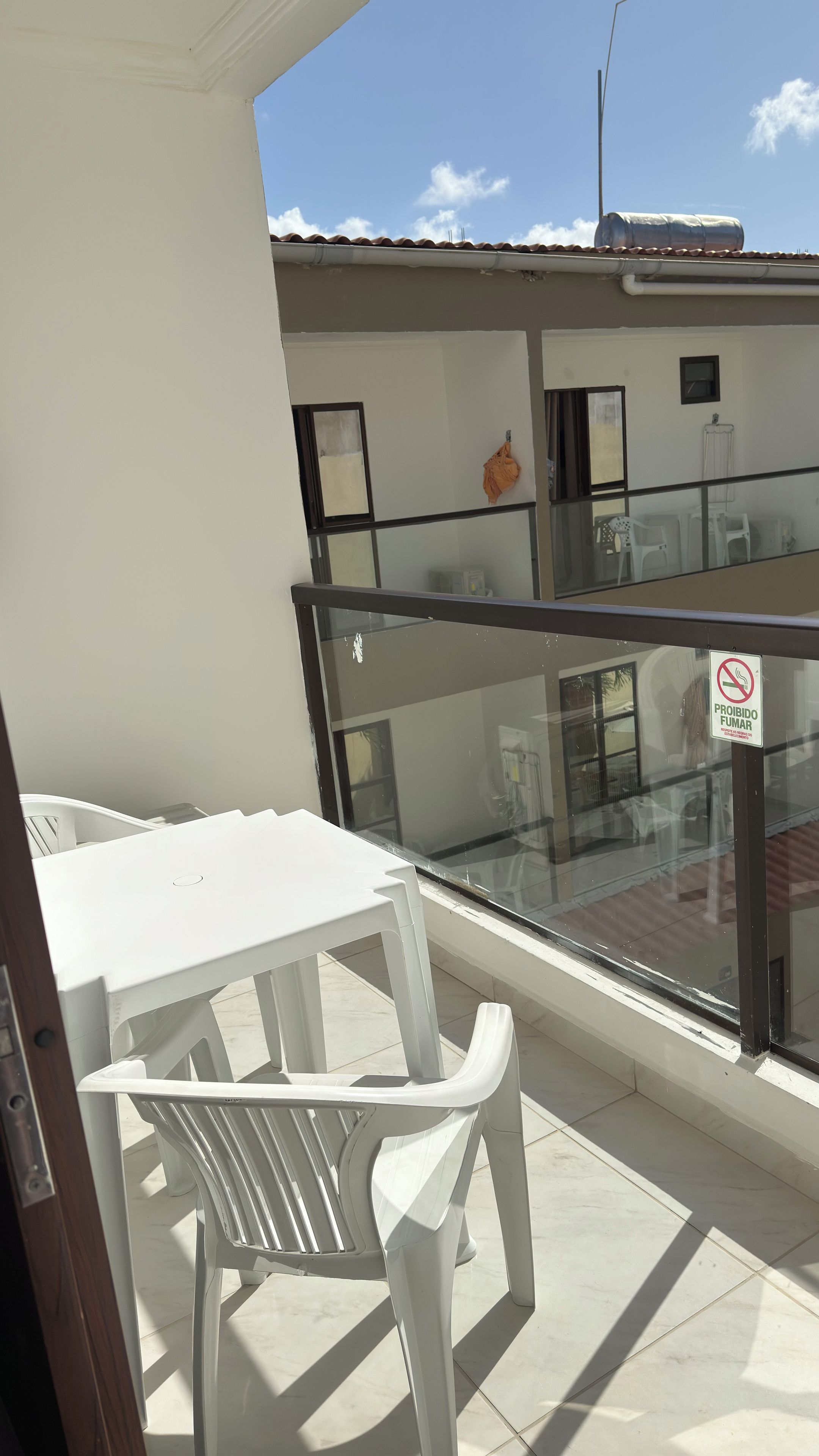 Un balcón con mesa y sillas blancas, barandilla de cristal y un edificio con tejado a la vista.