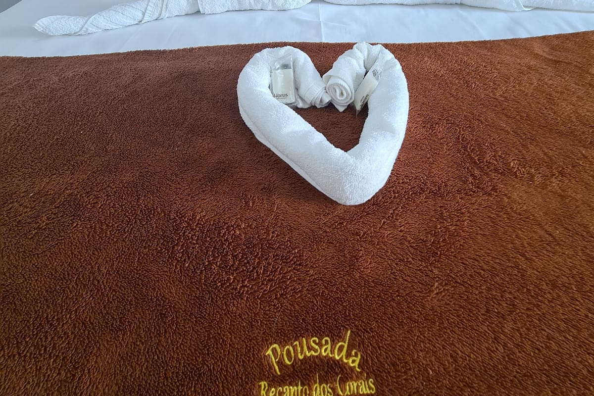 Ein Hotelzimmer mit einem Bett, das mit weißen Leinen bezogen ist, und einem braunen Teppich mit Struktur. Das Bett ist mit einer herzförmigen Handtuchanordnung und einem herzförmigen Handtuch auf dem Teppich dekoriert. Auf dem Teppich ist der Schriftzug "Pousada Recanto dos Corais" zu sehen.