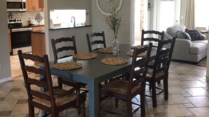 En matplats med ett blått bord och trästolar, ett kök med träskåp och en mikrovågsugn, och ett vardagsrum med en soffa och gardiner.