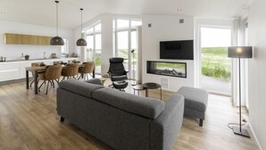 Ein modernes Wohnzimmer mit einem grauen Sofa, einem Essbereich mit Tisch und Stühlen, einem an der Wand befestigten Fernseher und einem großen Fenster mit Blick ins Freie.