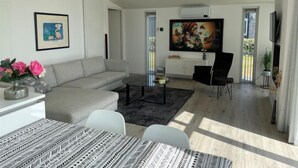 Ein modernes Wohnzimmer mit grauem Sofa, Couchtisch, Esstisch mit gemustertem Tischläufer, Fernseher und Bildern an den Wänden.