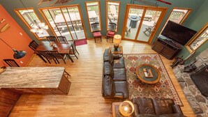 A spacious living room with a wooden floor, a stone fireplace, a leather sofa, a dining area with a wooden table and chairs, and a large TV. - Cozy Cove Retreat w/ Arcade & Fireplace (Oakland)