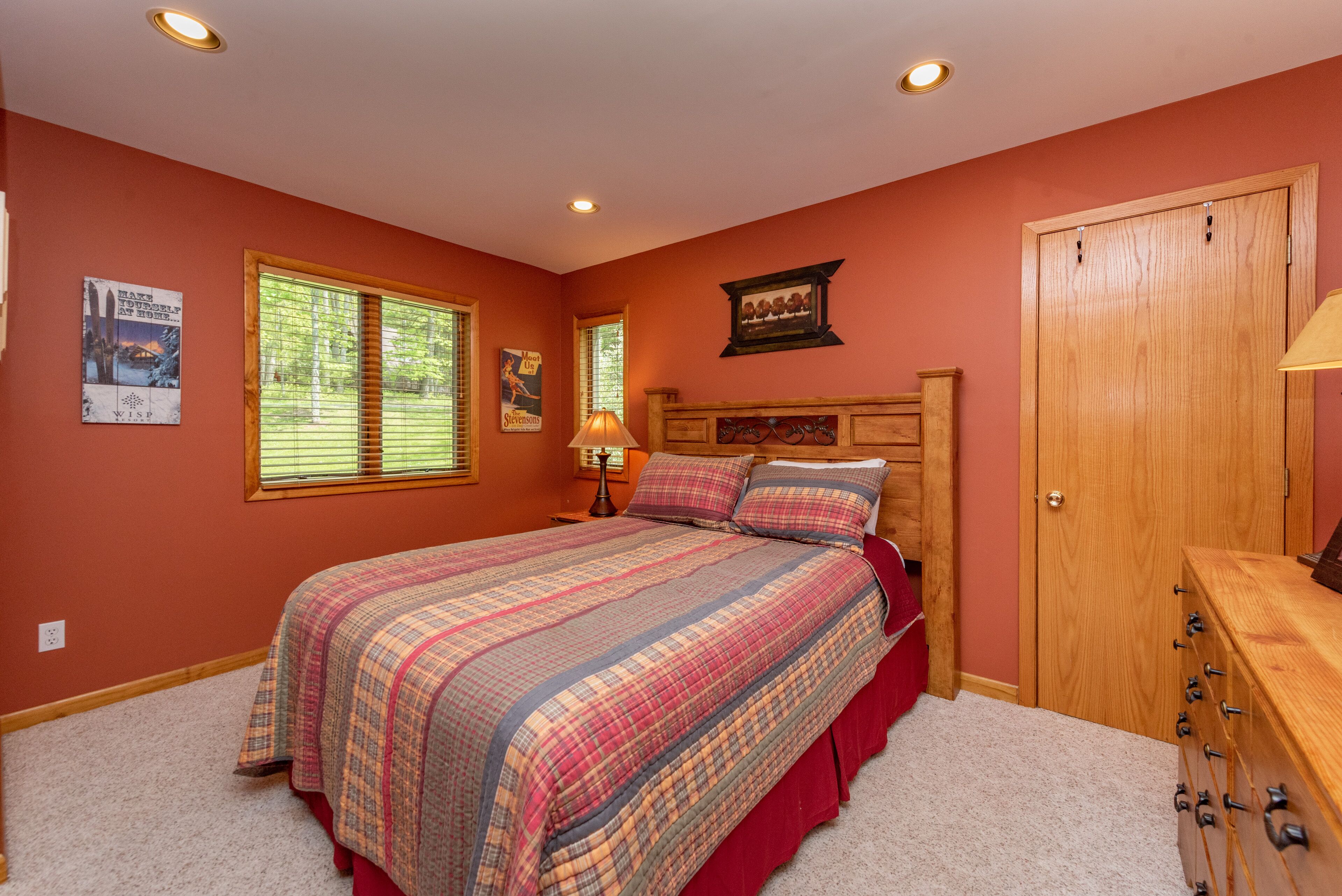 A bedroom with a large bed, a wooden dresser, a closet, and two windows with blinds.
