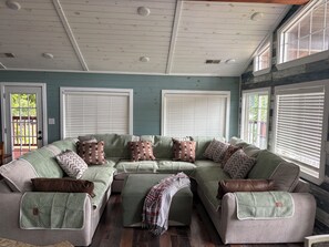 A cozy living room with a sectional sofa, a fireplace, and large windows with blinds.
