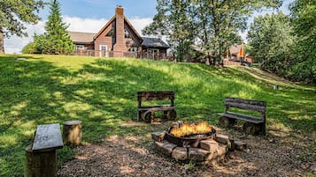 A rustic cabin with a fire pit, wooden benches, and a grassy hill.