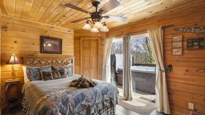 A wooden cabin bedroom with a bed, a ceiling fan, a framed picture, and a window with curtains. - Traditional Log Cabin Near Big Cedar, PVT Fire Pit, Hot Tub, Scenic Woods, Trails End, Free Tickets! (Ridgedale)