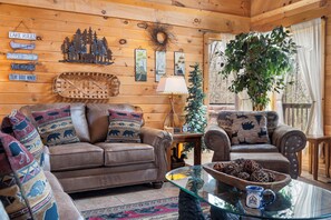 Ein gemütliches Cabin-Interieur mit einer Holzwand, einer Couch, Sesseln, einem Glastisch und einem dekorativen Kranz.
