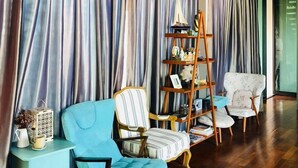 A room with a wooden floor, striped curtains, a blue sofa, a striped armchair, a wooden ladder shelf, and a small table with a lantern.