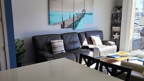 Un salon moderne avec un canapé en cuir noir, une table basse blanche et un grand tableau représentant une jetée.