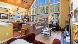 A spacious living room with a high ceiling, large windows, and a fireplace. - Moose Manor | Hot tub, Wifi, Fireplace, AC (Packwood)