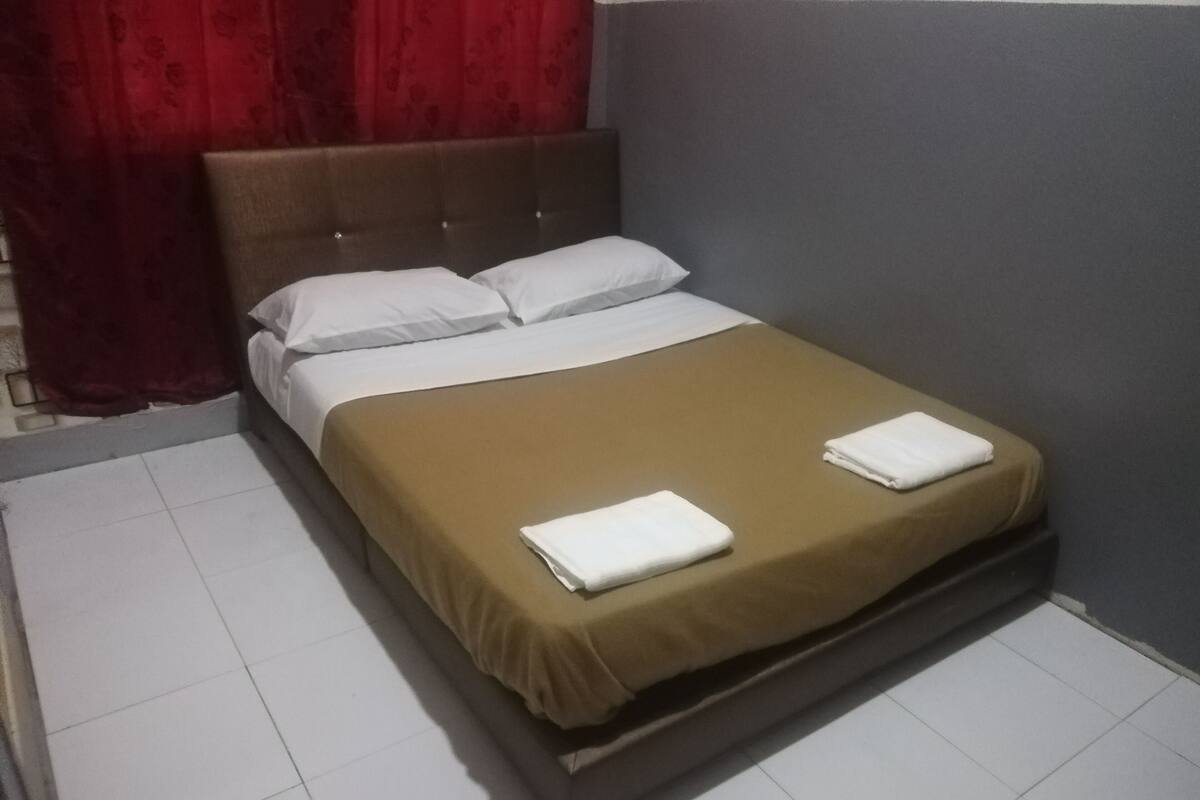 A single bed with a brown bedspread and white pillows in a room with a grey wall and red curtains.