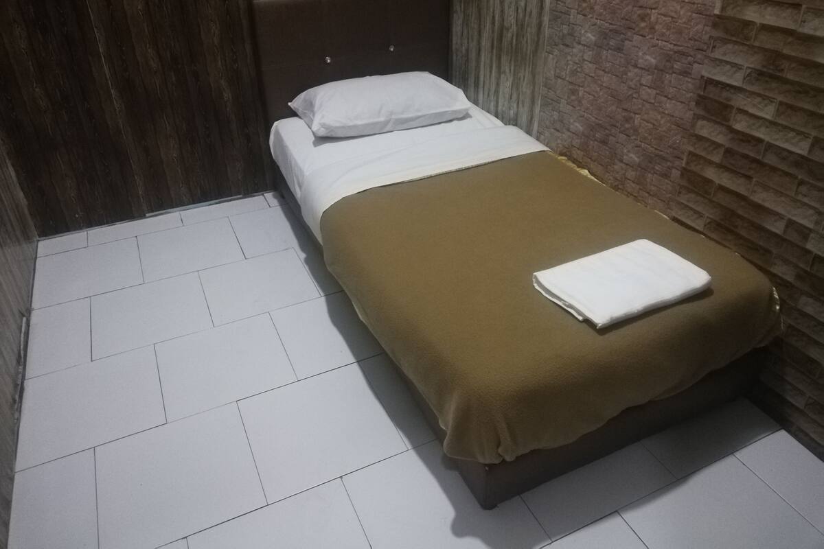 A single bed with a brown blanket and white pillow in a room with a tiled floor and a brick wall.