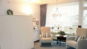 A room with a white wardrobe, two armchairs, a table with a lamp, and a window with sheer curtains. - App. Bölting 11 (Wenningstedt-Braderup)
