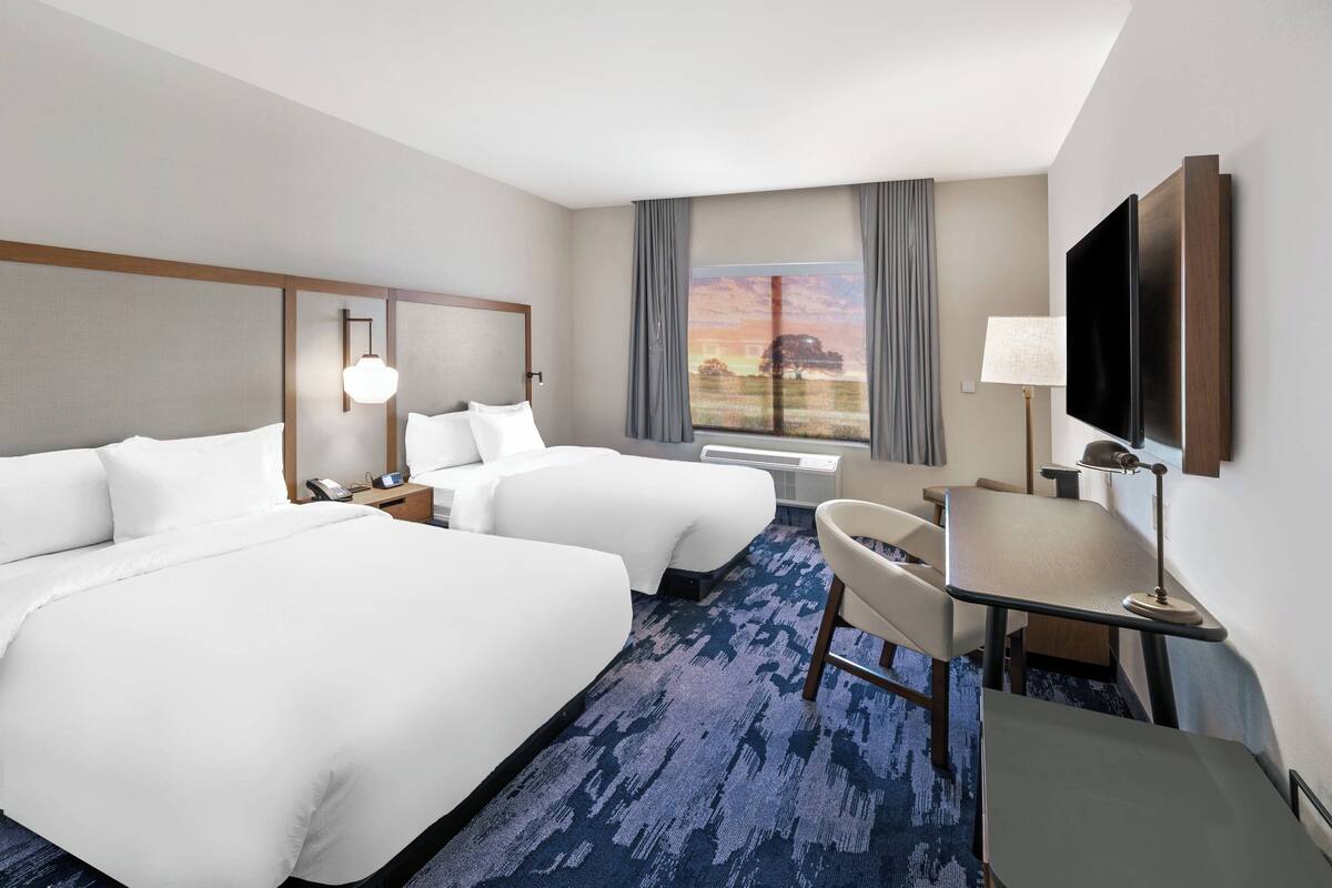 A hotel room with two beds, a desk, a chair, a TV, and a window with curtains.