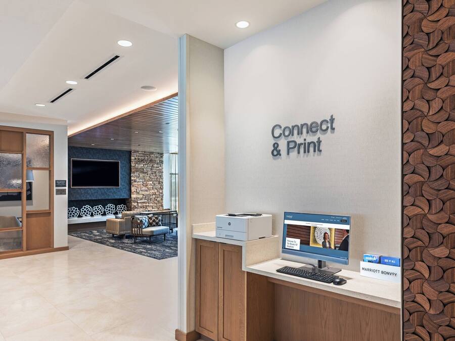 A hotel reception area with a 'Connect & Print' station, a seating area with a stone wall, and a large flat-screen TV.