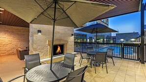 Un patio al aire libre con chimenea de piedra, mesas y sillas de metal, y sombrillas que dan sombra.