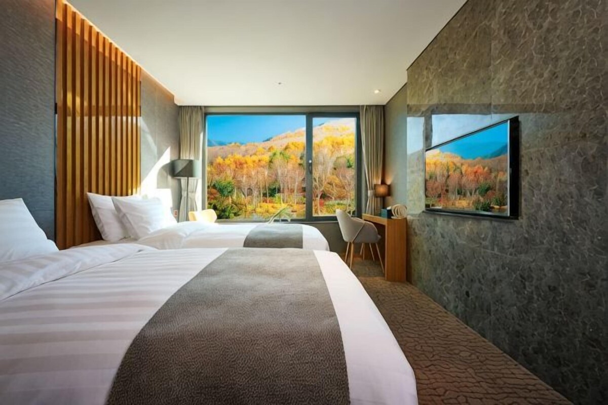 A hotel room with two beds, a desk, a chair, and a large window with a view of autumn foliage.