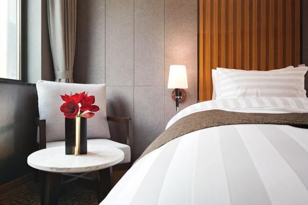 Habitación de hotel con cama, una mesa redonda con un florero y una lámpara de pared, además de un ventanal con cortinas.