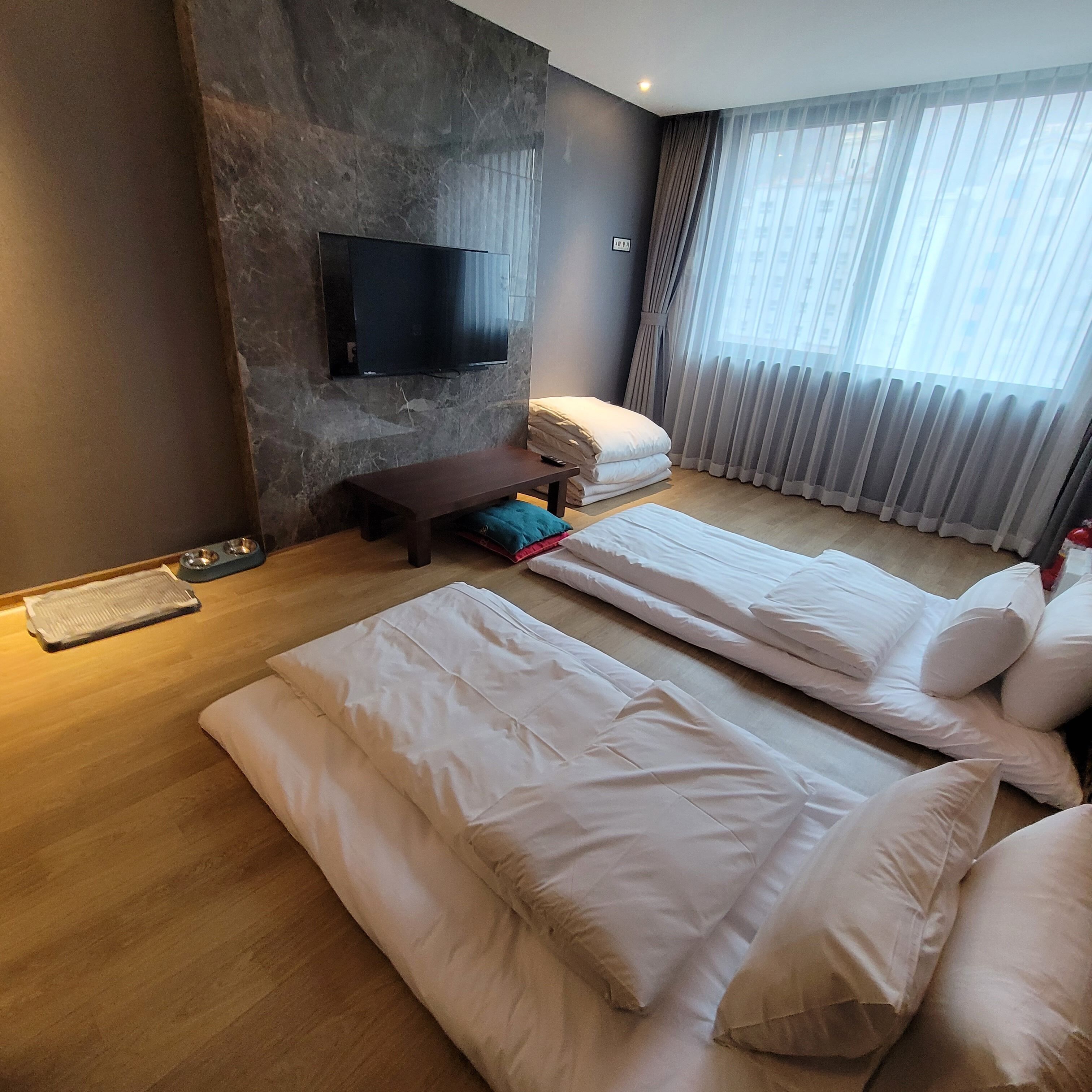 A modern hotel room with a flat-screen TV mounted on a textured wall, a wooden floor, a sofa bed with white bedding, a small table with a remote control, and a window with sheer curtains.
